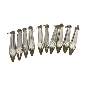 May include: A collection of clear glass chandelier prisms. Each prism is teardrop-shaped with a faceted design and a metallic silver-colored tip. Small metal loops are attached to the top for hanging. The prisms are arranged against a white background.