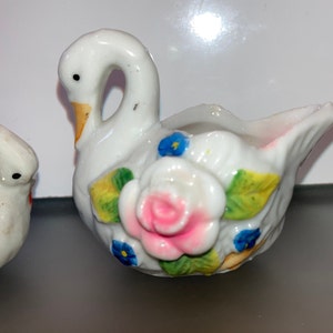 May include: A white porcelain swan figurine with a pink and white rose and blue flowers painted on the side. The swan has a yellow beak and is holding a small bowl.