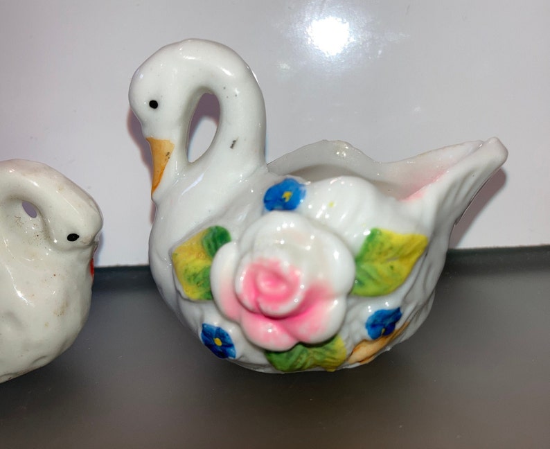 May include: A white porcelain swan figurine with a pink and white rose and blue flowers painted on the side. The swan has a yellow beak and is holding a small bowl.
