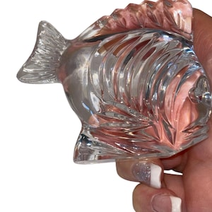 K&ouml;nnte beinhalten: Eine klare Glasfischfigur mit detaillierten Schuppen und Flossen. Der Fisch wird in einer Hand gehalten, was sein kompliziertes Design und seine reflektierenden Eigenschaften hervorhebt. Die Figur ist ein dekorativer Gegenstand.