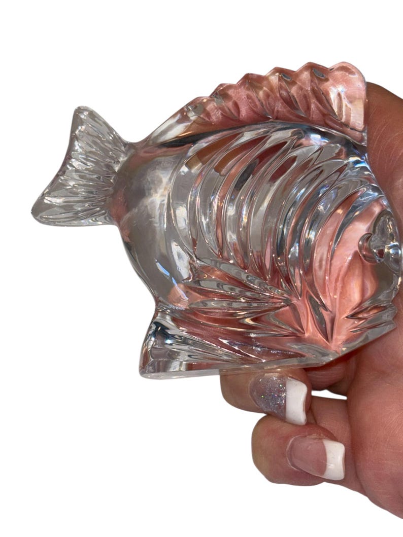 K&ouml;nnte beinhalten: Eine klare Glasfischfigur mit detaillierten Schuppen und Flossen. Der Fisch wird in einer Hand gehalten, was sein kompliziertes Design und seine reflektierenden Eigenschaften hervorhebt. Die Figur ist ein dekorativer Gegenstand.