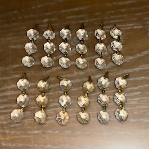 Pode incluir: Uma coleção de prismas de lustre de cristal. Cada prisma é facetado e conectado com ferragens de metal dourado. Os prismas são dispostos em fileiras verticais, criando uma exibição brilhante e elegante. O fundo é uma superfície de madeira quente.