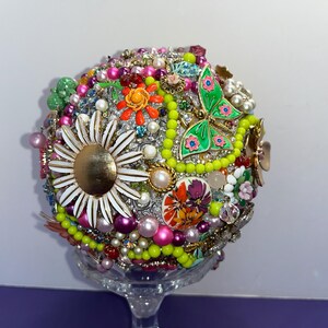 May include: A colorful sphere made of various vintage brooches, buttons, and beads. The sphere is decorated with flowers, butterflies, and other whimsical designs. The sphere is displayed on a clear glass stand.