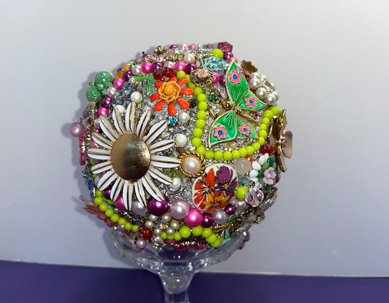 May include: A colorful sphere made of various vintage brooches, buttons, and beads. The sphere is decorated with flowers, butterflies, and other whimsical designs. The sphere is displayed on a clear glass stand.