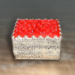 May include: A small, square, silver trinket box with a red floral design on the lid. The box has a decorative, ornate border around the lid.