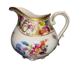 May include: White porcelain creamer with a gold rim and handle, adorned with colorful floral patterns. The creamer features a bulbous body and a curved spout. The floral design includes pink, blue, yellow, and orange flowers.