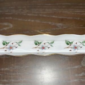 May include: White porcelain trinket dish with a wavy edge and gold trim. The dish is decorated with three white flowers with red centers and green leaves.