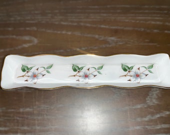 May include: White porcelain trinket dish with a wavy edge and gold trim. The dish is decorated with three white flowers with red centers and green leaves.