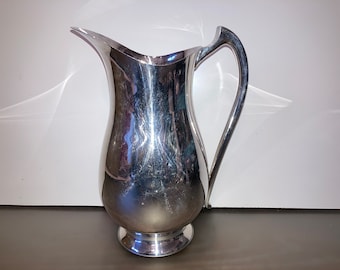 May include: A polished silver pitcher with a curved handle and a flared spout. The pitcher has a bulbous body and a circular base. The reflective surface shows slight scratches. This elegant pitcher is suitable for serving beverages.