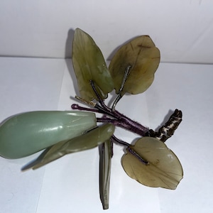 Puede incluir: Una escultura tallada en jade verde que representa un árbol con hojas y un solo fruto. El árbol está hecho de jade y tiene una base de madera marrón. Las hojas están talladas en un estilo realista y el fruto es una simple forma ovalada.