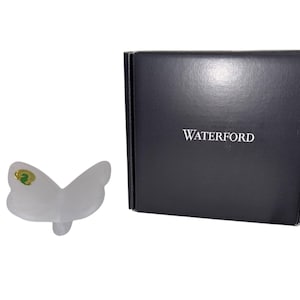 May include: A frosted glass butterfly figurine with a green and gold sticker, next to a black box with the word "WATERFORD" in white. The butterfly has a delicate, translucent appearance.