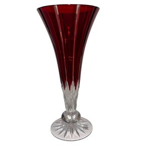 May include: A tall, elegant vase with a flared, ruby-red top and a clear, faceted base. The vase features vertical ribbing on the upper portion and a round, clear glass ball connecting the top to the base. The base is a starburst design.