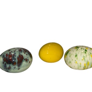 Könnte beinhalten: Drei dekorative Keramikeier. Eines ist türkis mit kastanienbraunen Flecken, das zweite ist leuchtend gelb und das dritte ist weiß mit grünen und gelben Sprenkeln. Die Eier sind oval und haben eine glänzende Oberfläche.