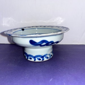 May include: A small, white ceramic bowl with a blue and white design. The bowl has a scalloped edge and a raised base. The design features a blue floral pattern.