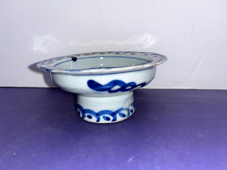 May include: A small, white ceramic bowl with a blue and white design. The bowl has a scalloped edge and a raised base. The design features a blue floral pattern.