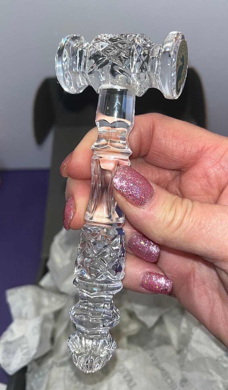 May include: A clear crystal gavel featuring intricate diamond-cut patterns on the head and handle. The head of the gavel displays a circular sticker with a seahorse emblem and the text Waterford Crystal. The piece has a faceted, reflective surface.