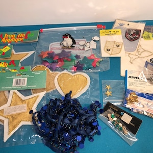 May include: Assortment of iron-on appliques, including stars, hearts, a penguin, and a coat of arms. There are also blue sequins, gold stars, and rhinestones.