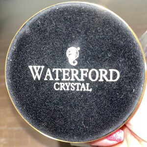 May include: A black circular base with a gold rim and the text "WATERFORD CRYSTAL" in white lettering.