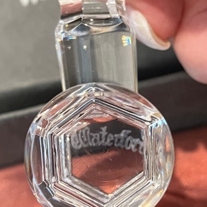 May include: A clear, faceted crystal glass stopper featuring a hexagonal top design. The word Waterford is etched into the center of the hexagonal face. The stopper has a multi-tiered, cylindrical neck with polished edges and reflective surfaces.