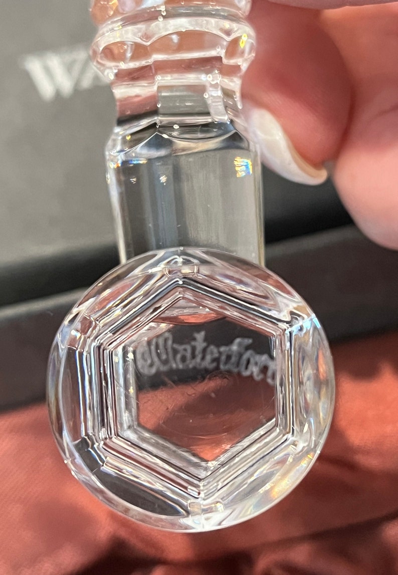May include: A clear, faceted crystal glass stopper featuring a hexagonal top design. The word Waterford is etched into the center of the hexagonal face. The stopper has a multi-tiered, cylindrical neck with polished edges and reflective surfaces.