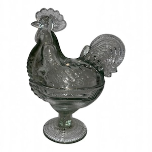 May include: A translucent gray pressed glass lidded dish shaped like a rooster. The piece features a textured surface with detailed feather patterns on the body, tail, and comb, standing on a circular pedestal base with a decorative rim.