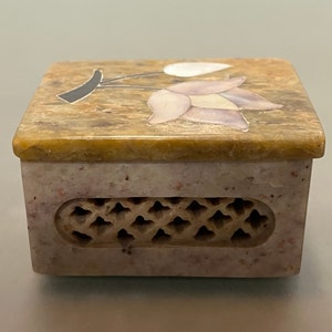 May include: A small, square, decorative box made of light brown and pink stone. The top of the box features a floral design with pink and white flowers and a black stem. The bottom of the box has a carved design.