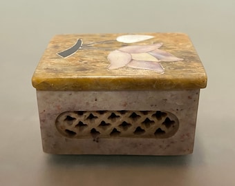 May include: A small, square, decorative box made of light brown and pink stone. The top of the box has a floral design with pink and white flowers and a black stem. The bottom of the box has a carved design.