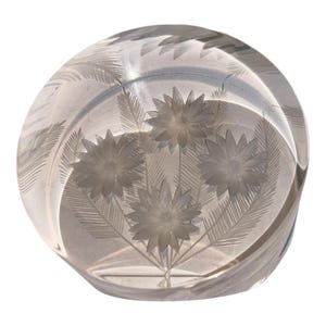 May include: A clear, spherical glass paperweight featuring an intricate, etched design of four blooming flowers with pointed petals and delicate, feathery leaves. The engraving is centered within the smooth, transparent glass orb.