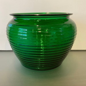 May include: A vibrant emerald green glass bowl featuring a rounded, bulbous shape with a series of horizontal, raised ridges encircling the exterior. The glass has a smooth, polished finish and a wide, open top with a slightly flared rim.