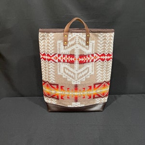 May include: A tall tote bag with a geometric pattern in shades of tan, red, and orange. The bag has a brown leather bottom and handles. The handles are attached with gold-colored rivets. The bag is made of a woven fabric.