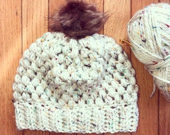 Op de afbeelding: Een witte gehaakte beanie met een bruine nep bont pompon. De beanie heeft een gespikkeld patroon met veelkleurige vlekken. Het garen is dik en de beanie is gemaakt met een textuur steek.