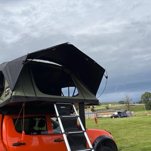 May include: An olive green and black rooftop tent mounted on an orange pickup truck. The tent features a black rainfly, a large open entrance, and a retractable metal ladder attached to the side. The structure is set against a cloudy sky.