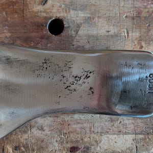 May include: A silver-colored axe head with the text "US98" stamped on the side. The axe head has a curved blade and a flat top. The axe head is lying on a wooden surface.