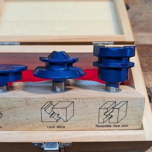 May include: A wooden box containing three blue router bits. The bits are labeled for drawer lock, lock mitre, and reversible glue joint applications. The box is open, revealing the tools and a red interior.