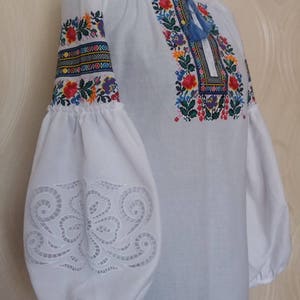 May include: White embroidered blouse with a floral pattern in shades of red, blue, yellow, and green. The sleeves have a lace-like design and the neckline is gathered.