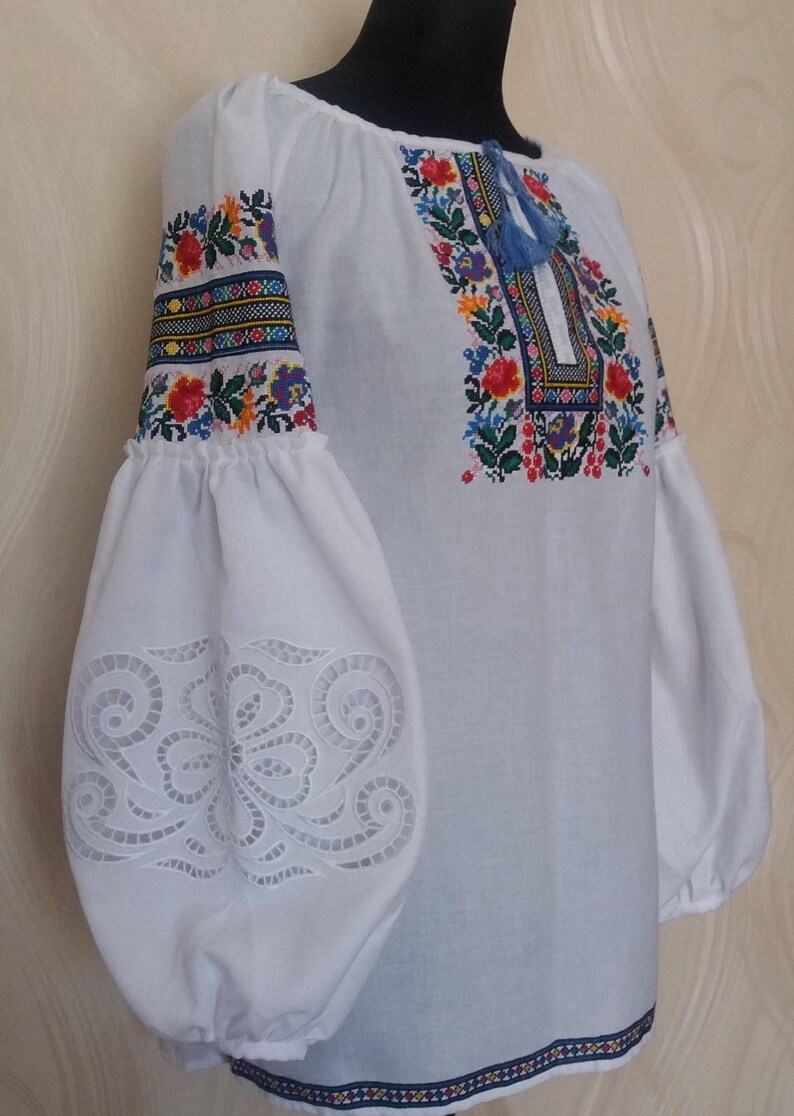May include: White embroidered blouse with a floral pattern in shades of red, blue, yellow, and green. The sleeves have a lace-like design and the neckline is gathered.