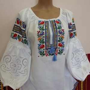 May include: White embroidered blouse with a floral pattern and blue trim. The sleeves have a lace design.