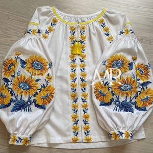 May include: White blouse with embroidered sunflowers in yellow and blue. The blouse has long, puffed sleeves and a round neckline with yellow trim. A yellow tassel hangs from the neckline. The embroidery is detailed and covers the sleeves and front.