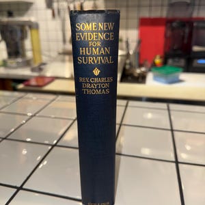May include: A vintage book with a navy blue cover and gold lettering. The title reads "SOME NEW EVIDENCE FOR HUMAN SURVIVAL" by Rev. Charles Drayton Thomas. The book is published by Collins.