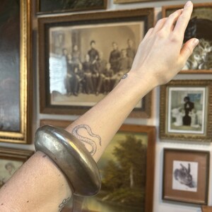 May include: A large, silver-toned metal bangle bracelet. The bracelet is oval-shaped and worn on the arm. The background features framed vintage artwork and photographs.