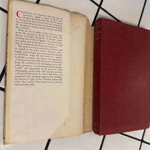 Puede incluir: Un libro abierto con texto y un libro cerrado, de color burdeos, titulado "The Secret of Dreams" por el Padre Meseguer. Las p&aacute;ginas del libro abierto son blanquecinas con texto negro. Los libros est&aacute;n sobre una superficie de baldosas blancas.