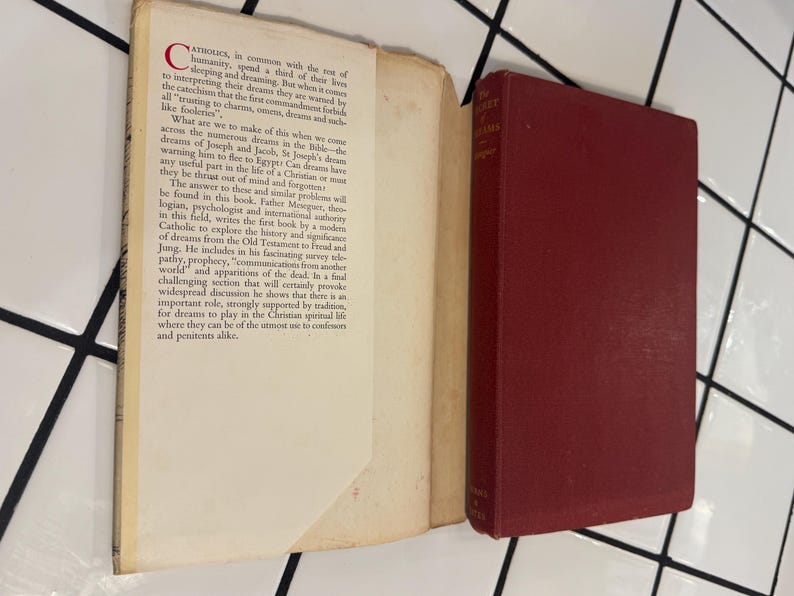 Puede incluir: Un libro abierto con texto y un libro cerrado, de color burdeos, titulado "The Secret of Dreams" por el Padre Meseguer. Las p&aacute;ginas del libro abierto son blanquecinas con texto negro. Los libros est&aacute;n sobre una superficie de baldosas blancas.