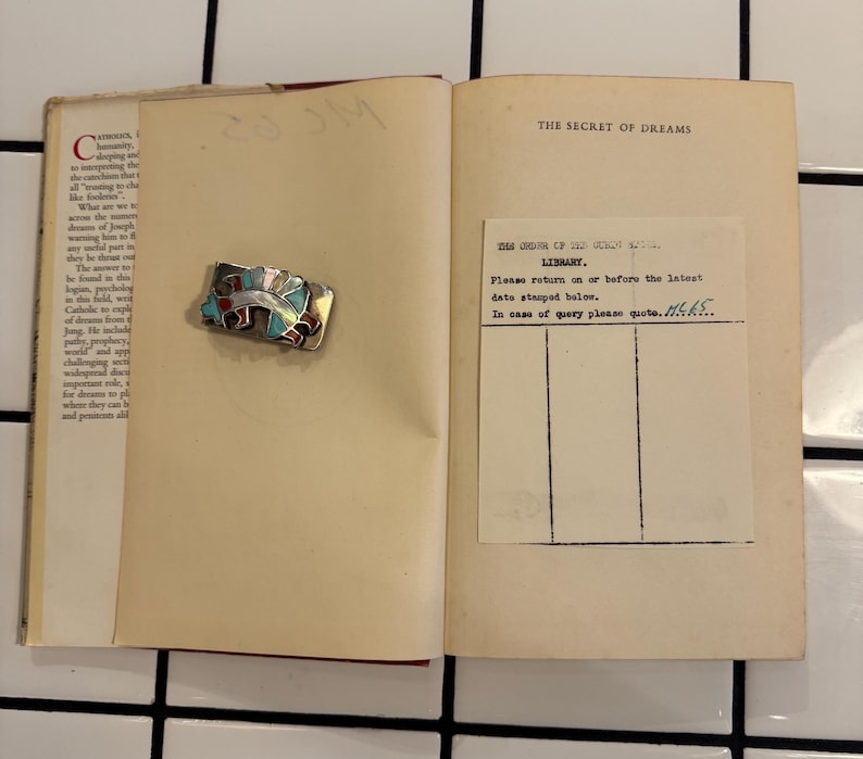 Puede incluir: Un libro abierto con el t&iacute;tulo "The Secret of Dreams" y una tarjeta de biblioteca. Un broche rectangular con detalles rojos, azules y plateados descansa en la p&aacute;gina izquierda. El libro est&aacute; sobre una superficie de baldosas blancas.