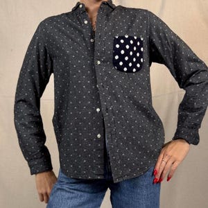 Könnte beinhalten: Ein dunkelgraues Button-Down-Hemd mit weißem Polka-Dot-Muster. Das Hemd hat eine marineblaue Tasche mit weißen Polka-Dots. Das langärmlige Hemd wird mit einer blauen Jeans kombiniert.