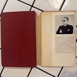 Puede incluir: Un libro abierto con una cubierta granate, que revela una p&aacute;gina con texto y un retrato en blanco y negro de un hombre con gafas. El texto incluye "THE AUTHOR" e informaci&oacute;n biogr&aacute;fica. El libro est&aacute; sobre una superficie de baldosas blancas.
