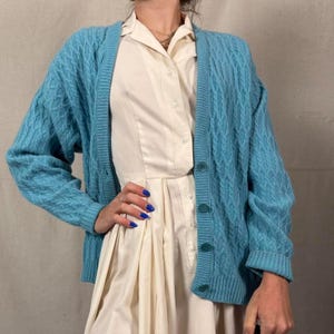 May include: A light blue cable knit cardigan with a button-up front. The cardigan is worn over a white collared dress. The sleeves are long, and the overall style is casual and comfortable. The cardigan is a solid color.