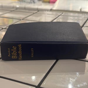 Puede incluir: Un libro azul marino "Pocket Bible Handbook" con letras doradas. El libro es rectangular y tiene el nombre "Halley" impreso en la portada. El libro está sobre una superficie de baldosas blancas.
