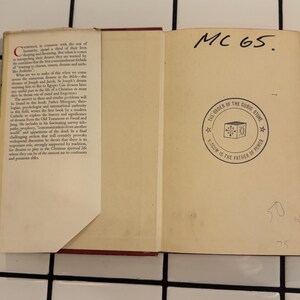 Puede incluir: Un libro abierto con lomo rojo y p&aacute;ginas blanquecinas. La p&aacute;gina izquierda tiene texto, mientras que la p&aacute;gina derecha presenta un emblema circular con texto y un dise&ntilde;o de cubo. Texto manuscrito "MC 65" en la parte superior.