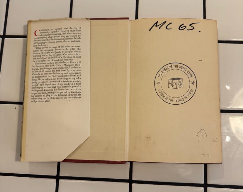 Puede incluir: Un libro abierto con lomo rojo y p&aacute;ginas blanquecinas. La p&aacute;gina izquierda tiene texto, mientras que la p&aacute;gina derecha presenta un emblema circular con texto y un dise&ntilde;o de cubo. Texto manuscrito "MC 65" en la parte superior.