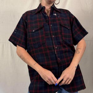 May include: A short-sleeved plaid shirt in navy blue, red, and green. The shirt has two chest pockets and a button-down front. The fabric appears to be a heavier weight, suitable for cooler weather. The shirt has a classic, vintage style.